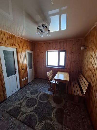 The image shows an interior room with wood-paneled walls and a white ceiling. The room contains a wooden table with two wooden benches on either side, a window, two white doors with frosted glass, a ceiling light fixture, a wall sconce, and a rug on the floor. The floor appears to be tiled with a patterned design. The scene depicts an empty room, likely a dining area or a common space. The lighting suggests it is daytime, with natural light coming from the window. The overall impression is of a simple, functional interior. The location in the city of Kara-Balta, Kyrgyzstan, is not discernible from the visual information provided in the image.