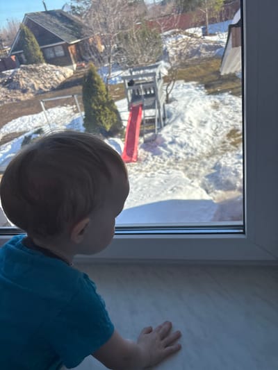 A young child, with light brown hair and wearing a short-sleeved blue top, is positioned with their back to the viewer, looking out of a white-framed window. The child's left hand rests flat on a light-colored, possibly marbled, windowsill. The scene outside the window, located in Shul'gino, Russia, depicts a residential backyard environment under bright daylight, indicative of late winter or early spring. The ground is partially covered in melting snow, interspersed with visible patches of brown grass and bare earth. A prominent wooden playset, featuring a red slide and a ladder, is situated in the middle ground. Various trees, including bare deciduous trees and green coniferous trees, are present. In the upper left background, a small, dark-roofed, brown house is visible behind a large pile of earth or debris. Distant fencing marks property boundaries.