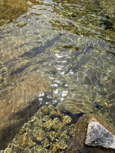 The image shows a close-up view of a shallow, clear riverbed with numerous rocks and pebbles. The water is disturbed, creating ripples and reflecting sunlight, which casts shimmering patterns on the submerged stones. The rocks are of various sizes and colors, ranging from dark gray to light brown and yellowish hues. Some larger, smoother stones are visible, as well as clusters of smaller, more angular pebbles. The bottom of the river appears to be sandy or silty in places, with some darker shadows suggesting deeper areas or underwater vegetation. The overall impression is one of a natural, undisturbed aquatic environment. The location is identified as a river in Hospental, Switzerland, suggesting a mountainous or alpine setting.