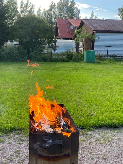 In Leipyasuo, Russia, a metal brazier stands on a gravel surface, with a vibrant fire burning inside. The flames are predominantly orange and yellow, consuming pieces of wood. The brazier is positioned in front of a lush green lawn, which extends to the background. Beyond the lawn, several houses with red tiled roofs are visible, nestled among trees. To the right of the houses, a green metal container is placed. The sky above is blue with scattered white clouds, suggesting fair weather. The scene captures an outdoor setting with a fire as the central focus.