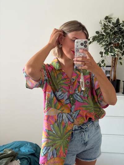The image shows a young woman taking a selfie in a bedroom. She is wearing a colorful floral print shirt and denim shorts. She has blonde hair and is wearing earrings and a ring. She is holding a phone in her hand, and a plant is visible in the background. There are also some clothes piled up on the floor. The image was taken inside a bedroom. There is no information available about the location of Cluses, France.