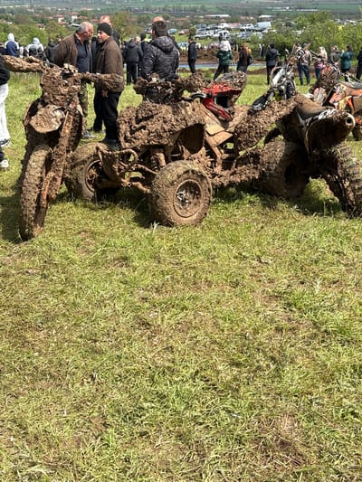 This image, rotated 90 degrees clockwise, depicts an outdoor scene during what appears to be an off-road event in Tsonevo, Bulgaria. The right side of the frame is a vibrant expanse of green and brown grass, while the left showcases a muddy area with several heavily encrusted off-road vehicles.

In the foreground and midground, multiple ATVs (quad bikes) and possibly dirt bikes are completely caked in thick, dark brown mud, evidence of strenuous activity in challenging terrain. One ATV in the foreground has a distinctive red helmet resting on its muddy chassis. The vehicles' tires and mechanical components are largely obscured by the clinging mud.

A group of adults is gathered around these vehicles, dressed in practical outdoor clothing, including jackets and beanies, suggesting cool weather conditions. They appear to be spectators or participants, observing the muddy machinery.

The background reveals a rural landscape with distant rolling hills or a valley, dotted with scattered buildings. A line of parked white vehicles, likely cars or vans, further in the background, indicates a larger organized gathering or event.

The scene is brightly lit by daylight, suggesting mid-day or early afternoon. The soft, even lighting implies an overcast sky, consistent with the muddy conditions. The heavy mud on the vehicles strongly indicates an off-road competition or rally, such as mud bogging. No visible text is discernible anywhere in the image.