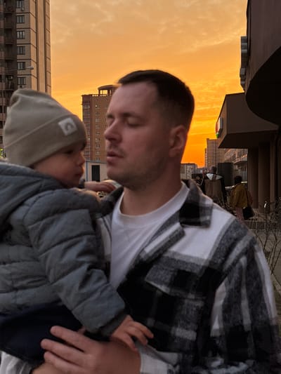 An adult male holds a young child in an urban environment at sunset in Gribovo, Russia. The male, central in the foreground, has short dark hair, wears a white t-shirt and a grey and black plaid flannel shirt, and has his eyes closed or directed downwards. He supports the child with his right arm. The child, positioned on the left, wears a grey beanie with a white rectangular logo and a grey puffer jacket, looking towards the male's face. The background features multiple multi-story residential buildings and commercial structures. The sky displays vibrant orange and yellow hues consistent with sunset, with light cloud cover. On the right, a building facade includes a partially visible sign with red and green elements. Two additional figures are visible in the mid-distance on the right, walking away from the camera on a paved surface alongside sparse, bare vegetation.