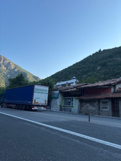 A blue semi-trailer truck with "EKOPRUT INTERNATIONAL TRANSPORT" written on its side is parked on the right side of a two-lane asphalt road. The truck has a license plate reading "BP 3324 BB". To the right of the truck are several buildings with red-tiled roofs. One building has a mural of people rafting on its wall, and a sign that says "RESPECT RAFTING TEAM". The road is bordered by a steep, forested mountainside on the left and the buildings on the right. The sky is clear and blue. The image was taken in Stara Kresna, Bulgaria.