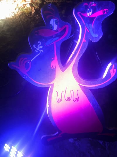 The image shows an illuminated, cartoonish, wood-cutout flamingo decoration in an outdoor setting. The flamingo is painted in blue and pink, with exaggerated facial features and stylized markings. A bright light source, likely a spotlight, is aimed at the decoration, casting a blue hue on the left side and a pink hue on the right side of the flamingo's body. The environment is dark, suggesting a nighttime setting, with some vegetation and possibly a dirt surface visible. Based on the location provided, this decoration may be found in a park or outdoor display in Auvers-Saint-Georges, France. There are no people present in the frame.