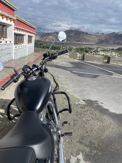A black motorcycle is parked on a paved surface overlooking a valley. The motorcycle is in the foreground, facing away from the viewer. The handlebars, fuel tank, and seat are visible. Behind the motorcycle, a red and yellow building with white trim is visible on the left. Prayer wheels are mounted on the building's exterior. The building is situated on a rise, overlooking a valley with a town or village and distant mountains. The sky is cloudy. The location is Thiksey, India.