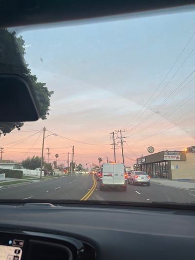 A view from inside a car shows a street scene at dusk. A white van and a silver sedan are in front, with a building partially visible on the right. Utility poles and lines stretch across the sky, which is tinted with pink and orange hues. The road has double yellow lines. The car's dashboard and windshield are in the foreground, partially obscuring the view.