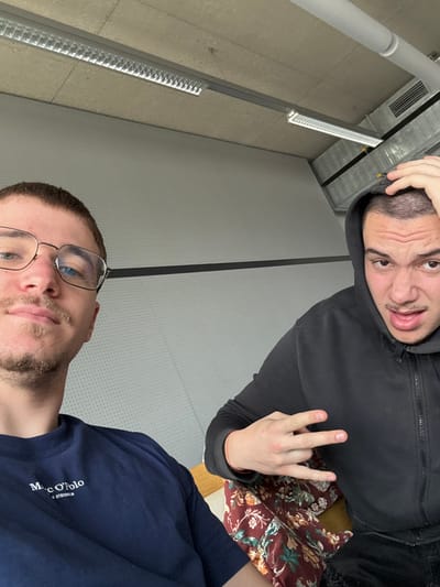 The image portrays two young adult males in an interior setting, understood to be located in Krems an der Donau, Austria.
The male on the left, positioned in the foreground and appearing to hold the camera, has short brown hair, blue eyes, a thin mustache, and stubble. He wears gold-rimmed eyeglasses and a dark blue t-shirt with a white "Marc O'Polo" logo. His expression is neutral as he looks directly at the camera.
The male on the right, partially visible, wears a dark hooded zip-up sweatshirt with the hood up, revealing short, dark, closely-cropped hair. His right hand is placed on his head, fingers spread. His left hand is raised, performing a hand gesture with the index, middle, and ring fingers extended while the thumb and pinky are bent. A thin red string bracelet is visible on his left wrist. He has an exaggerated facial expression with raised eyebrows, an open mouth, and visible teeth. He sits on a surface covered by a dark, floral-patterned fabric in shades of maroon, orange, and green.
The background features light grey perforated wall panels with a distinct horizontal black stripe. Above the wall, an exposed concrete ceiling is visible, along with multiple linear industrial fluorescent light fixtures and metallic ventilation ducts. The environment suggests a modern, functional, possibly public or institutional interior space.