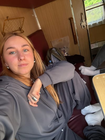 A first-person, overhead shot shows a woman lying on a burgundy-colored train seat in a compartment with wood paneled walls. She is wearing a gray tracksuit and silver earrings and rings. In the background, two sets of feet in white socks are visible under a wooden table. Other objects include a brown metal rack, a bag labeled "Rina", and a backpack. The location is assumed to be Радашковіцкі сельскі Савет, Belarus.