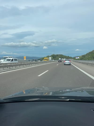 The image depicts a view from the driver's perspective of a highway. The road is paved with asphalt and has white lane markings. There is traffic on the highway, with several cars and vans visible in front and in other lanes. The vehicles include a silver hatchback, a yellow van, and several white vans. In the distance, there are trees and hills, suggesting a rural or suburban setting outside of a city. The sky is cloudy with a mix of grey and white clouds. There is a metal guardrail on the side of the highway. A yellow triangular sign with a black symbol is visible on the left side of the road. The dashboard of the car is visible in the foreground, reflecting the highway and sky. The scene is consistent with a highway in or near Gorno Varshilo, Bulgaria, given its general appearance of a European highway with surrounding nature.