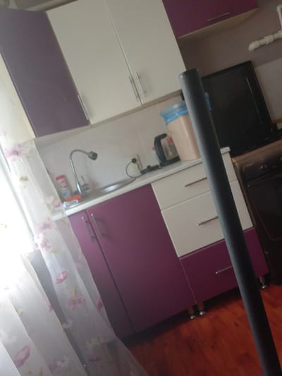 The image shows a kitchen with purple and white cabinets. On the counter, there is a sink, a faucet, a bottle of cleaning spray, and a red sponge. To the right of the sink, there is a white kettle and a beige bin with a blue lid. In the background, a dark television screen is visible. A dark grey pole runs vertically through the foreground of the image. The floor is made of polished wood. The room appears to be located in an apartment in Otradnaya, Russia.