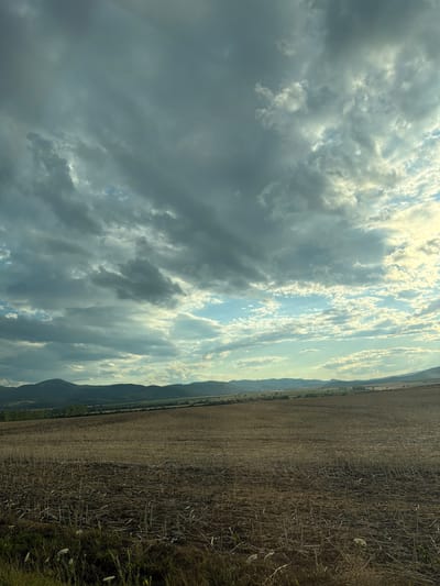 An expansive, low-angle shot captures a harvested field in the foreground, characterized by dry, stubbled stalks and patches of dried grass. Beyond the field, rolling hills recede into the distance, their slopes dotted with scattered trees and vegetation, suggesting a rural or semi-rural environment. The horizon is marked by a range of layered, undulating mountains, their dark silhouettes contrasting with the sky. The sky itself is dominated by dramatic clouds, a mix of dark, brooding storm clouds and lighter, scattered cumulus clouds illuminated by streaks of sunlight. The overall atmosphere is one of natural beauty, with a sense of approaching or receding weather. The location is identified as the city of Luhyňa, Slovakia. No people or specific actions are depicted.