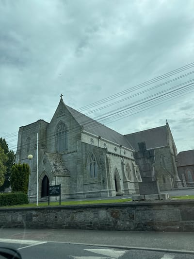 The image shows the exterior of St. Clare's Church in Carlow, Ireland. The church is constructed of grey stone with Gothic architectural elements, including pointed arch windows and a prominent cross on the roof. There is a sign in front of the church that reads "ST CLARE'S MASS TIMES" followed by a schedule of services. Power lines are visible overhead. The foreground shows a stone wall and a road with a crosswalk. The sky is overcast and cloudy.