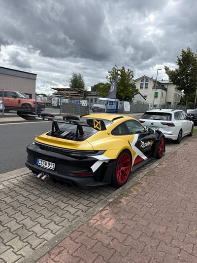 A heavily customized yellow and black Porsche 911 is parked on a multi-toned paver sidewalk adjacent to an asphalt road, located at Pfaffenberger Straße 21a, Hohenstein-Ernstthal, Germany. The vehicle features a large black rear wing with yellow endplates displaying a stylized "X" logo, red multi-spoke wheels, and a custom livery transitioning from yellow on the rear and upper body to black on the lower body and front. A prominent white "X" graphic with "SWdesign Karosseriebau & Fahrzeugtechnik" text is visible on the side. The German license plate reads "C SW 911". Two circular exhaust pipes protrude from the black rear diffuser.
In the background, a white Volkswagen Golf Variant (license plate "C PC 7372") is parked on the street. To the left, behind a chain-link fence, a peach-colored Land Rover Defender is partially visible in a commercial yard containing stacked wooden materials and multiple white vans. A light-colored industrial-style building and a multi-story building with a light facade and green lower section are present. A black flag displaying a white "SW" or "SW design" logo is mounted on a pole in the mid-ground. Green trees are interspersed, and the sky is overcast with dense grey clouds. No individuals are present in the scene.