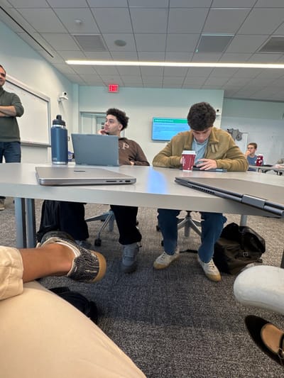 This image depicts an indoor scene within a classroom or meeting room located in Wellesley, United States. The environment features light-colored walls, a grey carpeted floor, and a suspended ceiling with square acoustic panels and fluorescent strip lighting. A prominent red "EXIT" sign is visible above a doorway on the back wall.

In the foreground, a light-grey rectangular table spans the width of the frame. On the left side of the table, a silver closed laptop (likely a MacBook) rests. Partially visible legs and feet belonging to an unseen person or the photographer are in the immediate foreground, showing light-colored pants and black-and-white patterned sneakers with tan soles. To the immediate right, another foot wearing a black shoe with a tan strap is visible.

Five individuals are visible. On the far left, a male with dark hair and a beard, wearing a dark green long-sleeved shirt and blue jeans, stands with his arms crossed, facing towards the right side of the frame, near a whiteboard with some writing. Seated at the table to his right is a male with dark curly hair, wearing a brown hooded sweatshirt with "GAP" visible on the chest and dark pants. He sits in an office chair, with a grey laptop (likely a Lenovo) open in front of him, and his left hand resting on his chin. A dark blue Yeti water bottle stands on the table to his left.

Further to the right, another male with dark wavy hair, wearing a light brown jacket over a light blue shirt and blue jeans, is seated. He holds a red and white "Costa Coffee" cup in his left hand while looking down at a dark tablet or laptop with a stylus/pen on the table in front of him. A dark backpack rests on the floor beneath his seat. Behind him, on the far right, a male with short dark hair, wearing a blue shirt, is partially visible, looking towards the front of the room. A large wall-mounted display screen on the back wall shows text including "Political and Socio-Economic." A white dome-shaped security camera is mounted on the wall above the whiteboard on the left.