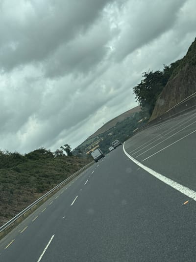 A white van and two cars are driving on a wet asphalt road with white lane markings. The road is bordered by a metal guardrail on the left and a steep, vegetated embankment on the right. The sky is overcast with dark, heavy clouds. The scene appears to be a highway or major road in a rural or semi-rural area. The image is tilted, suggesting it was taken from a moving vehicle. The location is likely County Wexford, Ireland, possibly on the N25 road near New Ross, given the commonality of such landscapes in the region.