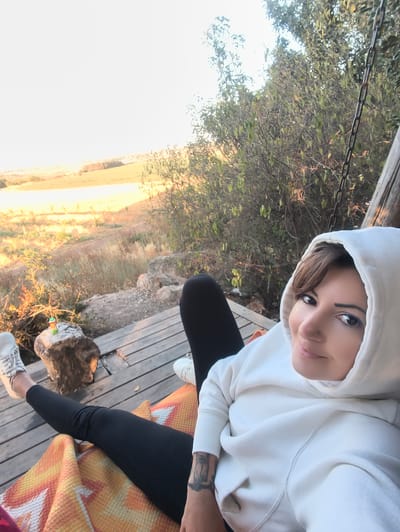 The image depicts a woman seated on a wooden deck in Shaalvim, Israel, looking directly at the camera with a slight smile. She has brown hair peeking from under a white hooded sweatshirt, black leggings, and white sneakers. A small nose piercing is visible, along with dark eyeliner and a tattoo on her left forearm. Her lower body and legs are covered by an orange, yellow, and white patterned blanket. To her left, a large rock on the deck holds a small, colorful cylindrical object. The deck is surrounded by natural rock formations and dry vegetation. In the background, a vast landscape of fields and hills extends under a bright sky, illuminated by warm, golden light. A metal chain hangs vertically on the right side of the frame, connected to a wooden post, suggesting a rustic outdoor structure.
