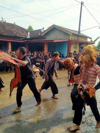 Men perform a street dance in front of a crowd of onlookers, their bare feet reflected in a wet road. One dancer holds a red, yellow, and black prop; another wears a shaggy mask and striped shirt. The crowd, mostly in dark clothing, watches from the sidewalk. A building with a tiled roof and blue exterior sits behind them, with utility poles and wires visible in the sky.