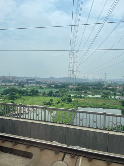The image captures a view from a train, looking out over a landscape in Dongguan, China. In the foreground, train tracks are visible, indicating the perspective. A concrete bridge railing with decorative metalwork separates the train from the scene beyond. The middle ground features lush green fields and bodies of water, suggesting a semi-rural or developing area. In the background, a city skyline is apparent, with numerous high-voltage power line towers stretching across the frame, a common sight in industrial or developing regions of China. The sky is overcast with clouds.