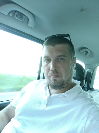 The image shows a man in the back seat of a car, looking directly at the camera. He has short brown hair, a light beard, and is wearing a white polo shirt. Sunglasses are perched on his head. He has a Bluetooth earbud in his right ear. The background shows the interior of the car, including the ceiling with a grab handle and light, and a window with a blurred view of a green, outdoor landscape, suggesting daytime travel. The lighting is bright, indicating it's likely a sunny day. The man's expression is neutral. There is no visible text.