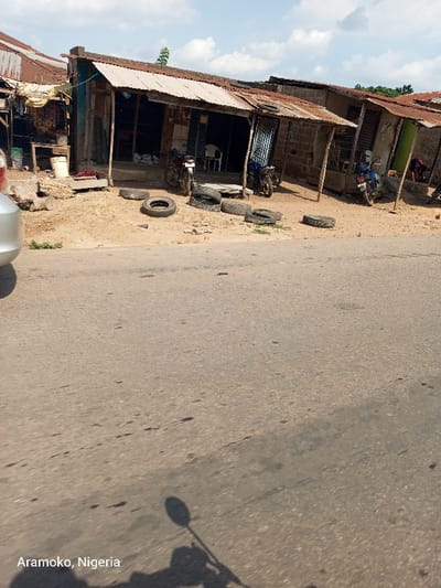 The image shows a street scene in Ilobu, Nigeria, with a row of shops along the side of a paved road. The shops are made of brick and concrete and have corrugated metal roofs. There are several motorcycles parked in front of the shops. The ground in front of the shops is covered with sand and there are several tires scattered about. The road is dusty and the sun is shining brightly. There is a shadow of a motorcycle in the foreground of the image. The coordinates provided locate the scene in Ilobu, Nigeria.