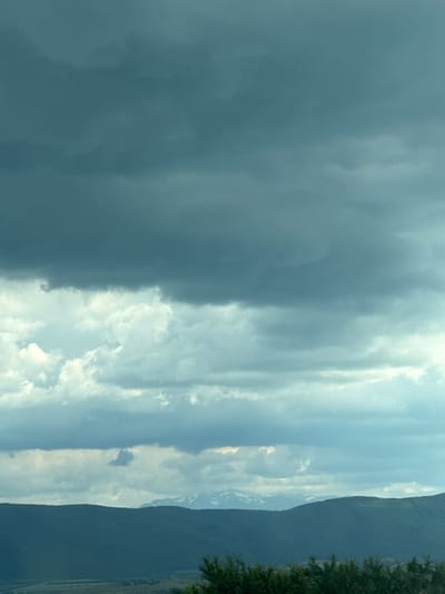A wide-angle, low-angle shot captures a dramatic sky filled with various cloud formations over a mountainous landscape in Rajana, Bulgaria. The upper portion of the image is dominated by dark, heavy clouds, tinged with a cool, blue-grey hue, suggesting overcast or stormy weather. Below these, lighter, wispy clouds drift, catching more illumination, creating a sense of depth and layering.
The horizon is defined by a series of dark green, undulating hills and mountains. In the distance, the peaks of higher mountains are visible, their summits capped with snow or frost, stark white against the muted landscape. The foreground shows a line of dense, dark green trees, their foliage providing a textured border to the scene. Below the distant mountains, a valley or plain is hinted at, with some lighter patches suggesting fields or sparse vegetation, though details are obscured by distance and the overall low light.
The environment appears natural and undeveloped, with no visible human activity, structures, or modern elements. The atmosphere is serene yet somewhat imposing due to the heavy cloud cover. The lighting is diffuse, casting no harsh shadows. The image is a landscape photography piece focused on atmospheric conditions and geological features.