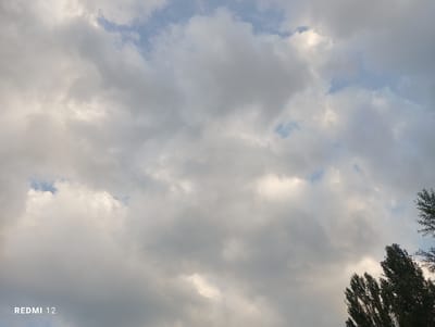 This is a photograph of a cloudy sky over Semetey, Kyrgyzstan. The image is dominated by gray and white clouds, with patches of blue sky visible. In the lower right corner, the tops of dark green trees are visible, with some branches extending into the frame. A watermark reading "REDMI 12" is present in the lower left corner of the image. The scene appears to be outdoors, with no people or specific landmarks of Semetey identifiable.