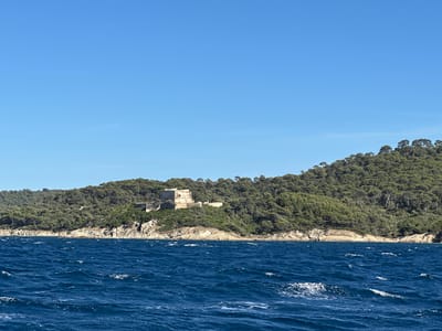 A still, eye-level, wide shot captures a panoramic view of the coastline of the Île du Levant, part of the Îles d'Hyères, off the coast of Hyères, France. The foreground is dominated by the deep blue, choppy waters of the sea, with white foam cresting the waves. The midground features a rocky coastline bordering a densely forested hillside. Perched atop the hill, a light-colored, weathered stone structure, possibly a fort or watchtower, is visible amongst the trees. The background is a clear, bright blue sky. No people are present in the image.