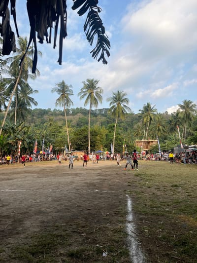 The image depicts an outdoor soccer match in Desa Batu Layar, Indonesia, under a partially cloudy blue sky. The foreground consists of a dirt playing field with sparse grass and a prominent white line marked vertically on the right side. In the midground, approximately 12-15 male players are engaged in a soccer game. One player in a light gray shirt and black shorts is positioned near a white soccer ball in the right-center, with another player in a dark red shirt and black shorts nearby. Other players wear red, white, or various colored shirts and shorts, distributed across the field.
A large crowd of spectators, numbering well over a hundred, lines the perimeter of the field, observing the game. Behind the spectators, on both the left and right sides, are various temporary structures including tents, canopies, and vendor stalls, adorned with colorful flags, some resembling the Indonesian flag. On the far right, a multi-level wooden scaffolding-like structure is visible. Numerous tall palm trees are scattered throughout the scene, particularly along the field edges and in the background. Lush green hills covered in dense tropical vegetation form the distant background. The lighting suggests daytime, possibly late afternoon. Overhanging, dark, dried banana leaves are visible in the upper foreground.