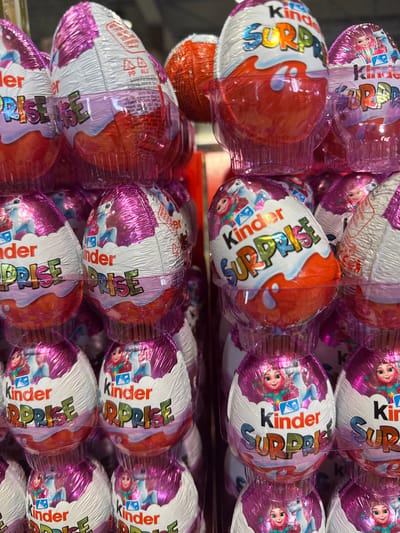 The image shows a close-up of a stack of Kinder Surprise eggs. The eggs are individually wrapped in foil, with half of each egg being white and the other half being either red or purple. The brand name "Kinder Surprise" is prominently displayed on the packaging. The environment suggests the image was taken at the city of Darien, in the United States of America.