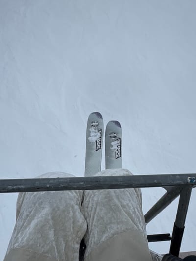 Overhead shot of skis and pants from a chairlift. The skis, branded K2, are gray with snow clinging to them. Thick, ivory-colored ski pants cover the skier's legs. A metal frame of the chairlift bisects the image, providing a partial view of the chair's structure. A vast expanse of snow is seen beneath and around the skis, filling a large portion of the frame.