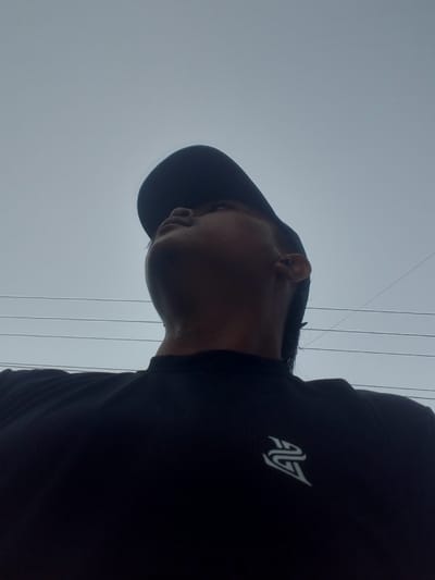 The image, taken from a low angle, shows the upper body of a person wearing a black t-shirt with a white logo. The person is also wearing a dark baseball cap, and is looking upwards. Their ear is visible and appears to have an earring. In the background, there are several horizontal power lines against a grey, overcast sky. The image does not contain any specific visual cues that would indicate the location is Kubang Jaya, Indonesia.