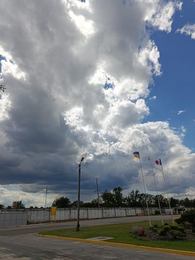 The media file shows an outdoor scene featuring a dramatic sky with a mix of dark, stormy clouds and bright, sunlit clouds against a vibrant blue sky. In the foreground, there is a paved road with yellow markings and a grassy area. A tall, concrete wall runs horizontally across the middle ground, with trees and some industrial structures visible beyond it.

Prominently featured are three flagpoles flying flags. One flag is blue and yellow, representing Ukraine. Another flag is white with a symbol, possibly a crest or emblem. The third flag is red, white, and blue, likely the French flag. The flags are positioned in the mid-ground, against the backdrop of the sky and the industrial setting.

A lamppost stands to the left of the flags, and power lines are visible against the darker clouds. The overall impression is a landscape of Ukraine, possibly on the outskirts of a town or near an industrial area, under a dynamic and weather-beaten sky. The contrast between the bright and dark clouds suggests a transition in weather, possibly a storm approaching or passing. The time of day appears to be daytime, given the light and shadows. There is no visible text in the media file, except for possibly very small markings on the wall or road that are not discernible.