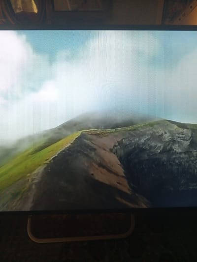 The original image is a photograph of a digital display, exhibiting visible horizontal scanlines across the entire screen. The display presents a mountainous landscape, purportedly located in the city of Puchezh, Russia. The depicted scene features a prominent ridge that sharply divides a vibrant green, grass-covered slope on the left from a dark, rugged, rocky terrain on the right. Dense mist and clouds partially obscure the upper portions of the mountains and the horizon, blending into a pale blue sky and indicating an overcast atmosphere. No individuals, animals, or artificial structures are discernible within the displayed landscape. The photograph's foreground includes the bottom bezel of the digital display, a light-colored curved support element beneath it, and a dark, textured surface. The upper part of the photograph shows reflections from the surrounding environment.