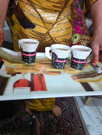 The image shows a white serving tray with three paper cups of coffee. The cups have a black and pink design with coffee beans and the word "COFFEE." The tray is placed on a patterned rug. In the background, a person wearing a yellow and brown patterned garment is partially visible, holding the tray. To the right of the tray, a hand with red nail polish is reaching for a cup. The scene appears to be indoors, as suggested by the rug and the lighting. The overall impression is one of hospitality, with coffee being served. There is no information in the image to suggest the location is Kawo, Nigeria.
