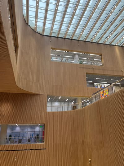 The image captures the multi-story interior of a modern building in Yanqiao, China. The dominant architectural feature is extensive light-colored vertical wood slat paneling covering both curved and flat walls, creating a warm, textured aesthetic. A large, sloped glass skylight forms the ceiling, supported by a grid structure and illuminated by integrated linear lights.
Multiple levels of the building are visible through large rectangular openings cut into the wood paneling. On the uppermost visible floor, an open, brightly lit space with a white ceiling, overhead fluorescent lighting, and a prominent white cylindrical support column is apparent, framed by glass railings.
On the mid-right side of the image, an escalator with a glass balustrade and a metal handrail ascends to an upper floor. Adjacent to the escalator, a decorative wall panel displays a grid of approximately thirty circular, multi-colored elements. Partially visible Chinese characters are present on a sign nearby.
In the lower-left portion, a rectangular internal window reveals a modern seating area. This area contains several contemporary armchairs with small tables and potted plants. A green emergency exit sign with white pictograms is mounted above this window.
No individuals are clearly discernible in the foreground or mid-ground, and any distant figures within the visible interior spaces are indistinct. The environment is meticulously maintained, well-lit by both natural light from the skylight and artificial illumination, and exhibits a clean, contemporary design.