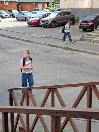 The image shows a woman walking on a sidewalk next to a street in Metkomelelo, Russia. She is wearing a striped shirt, blue jeans, and a backpack. She is looking down at her phone, which she is holding in her hands. The woman is walking towards the viewer.
In the background, there are several cars parked along the street. The buildings in the background appear to be made of brick. The sky is overcast, suggesting it may be a cloudy day. The time of day is difficult to determine precisely, but it appears to be daytime.
The scene is an urban environment, possibly a residential area or a commercial street. The woman appears to be focused on her phone, and her expression is neutral. There is a wooden fence or railing in the foreground, which is partially obscuring the view.
Visible text on one of the cars reads "K742AT 150 RUS".