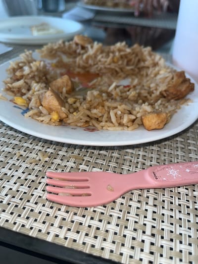 The image shows a plate of chicken fried rice next to a pink, child-sized fork on a beige woven placemat. The plate is mostly full of rice with visible pieces of chicken and egg. There are a few stray rice grains on the placemat. The photo appears to have been taken indoors. The location in La Victoria, Peru cannot be determined from the image.