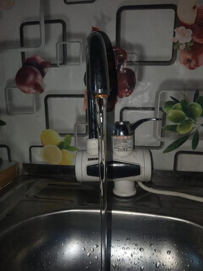 A close-up, eye-level shot shows a chrome faucet with water flowing into a stainless steel sink. Behind the faucet, a white device with a small lever is attached to the wall, which is decorated with a tiled pattern featuring images of pomegranates, lemons, and olives. The sink is wet, with water droplets visible on its surface. The location is the city of Ақадыр, Kazakhstan.