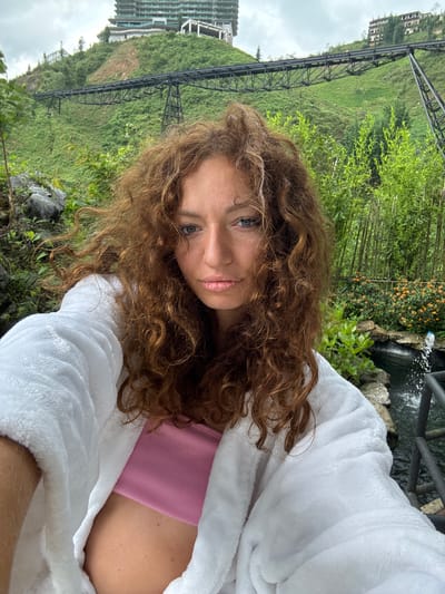 A woman with reddish-brown curly hair is seen in an eye-level medium shot in Cat Cat Village, Vietnam. She is wearing a pink top and a white bathrobe. The background includes lush greenery, a hillside, and a railway structure. A building is visible in the background on the left side. A small waterfall and some bamboo are visible behind the woman.