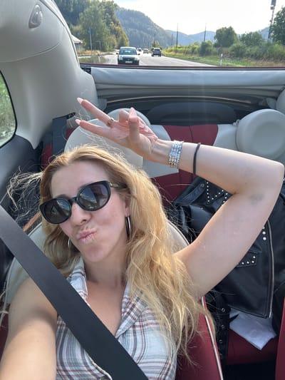 A blonde woman is taking a selfie while riding in a convertible. She is wearing sunglasses, a plaid shirt, and a bracelet. She is making a peace sign with her fingers. The car is driving on a road with trees and mountains in the background. The location is the B33 near Hausach, Germany.