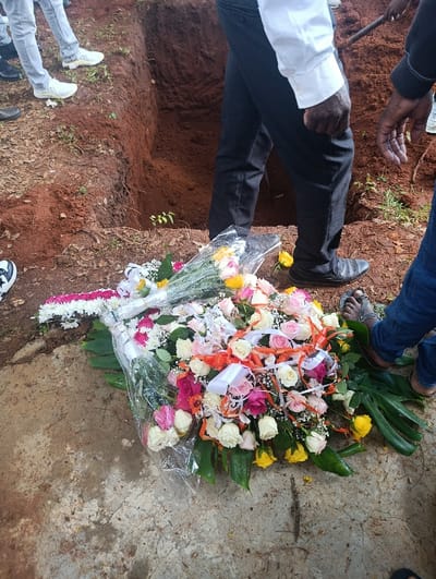 The scene depicts an outdoor burial site in Muthetheni ward, Kenya. A freshly dug rectangular grave, approximately two meters deep in reddish-brown soil, is visible in the mid-ground. Mounds of displaced reddish-brown soil surround the grave opening. Several individuals are present, with only their lower bodies and extremities visible. On the left, a person wears light gray jeans and white sneakers. In the center-right, a person wearing black trousers and a white shirt stands with black dress shoes near the grave's edge. To the far right, an individual in blue jeans wears dark sandals. In the upper right background, a dark-colored tool handle, consistent with a shovel, is partially visible, indicating active grave work. In the foreground, resting on a light gray, cracked concrete-like surface, is a large floral arrangement. It features an assortment of pink, white, yellow, and orange roses, interspersed with baby's breath and broad green foliage, partially encased in clear plastic film. White and orange ribbons adorn the arrangement. A smaller, elongated floral arrangement composed of pink and white flowers is partially visible behind the main one.