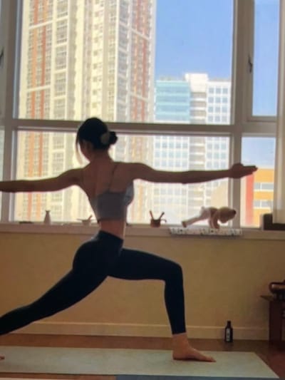 In a room with a large window overlooking an urban landscape of tall buildings, a person is performing a yoga pose on a mat. The person, seen from the back and side, is wearing a gray sports bra and black leggings. Their arms are extended to the sides, and their legs are in a wide stance, with one leg bent and the other extended. The window reveals a bright, clear sky and multiple high-rise buildings in various colors, suggesting a city environment. On the windowsill, there are a few small decorative objects, including what appears to be a stuffed animal. A dark bottle is also visible on the floor near the window. The overall scene suggests a domestic setting where the person is engaged in a personal fitness activity.