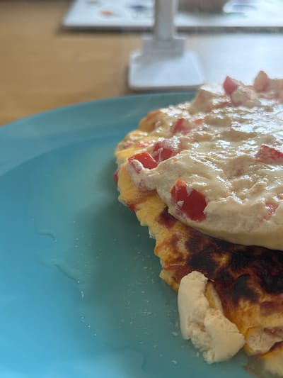A close-up shot focuses on a serving of food, likely an omelet, presented on a vibrant blue plate. The omelet appears to be filled with small diced red pieces, possibly bell peppers or tomatoes, and is topped with a creamy white sauce that also contains red flecks. The underside of the omelet shows signs of being cooked, with some darker, slightly charred sections. A few droplets of liquid are visible on the surface of the blue plate to the left of the food. The background is blurred, suggesting an indoor setting with a wooden table and some indistinct objects, including what looks like a white stand or holder. The scene is indoors, and no specific location in Kirtorf, Germany is depicted.