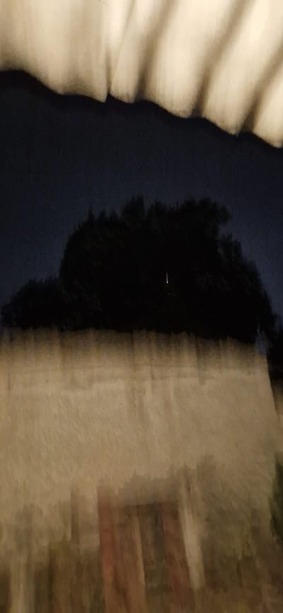 The image shows a blurry, dark landscape with a hint of a beige object at the top. The darkness suggests it might be night or a very shaded area. There's a silhouette of trees against the dark sky. The lower portion of the image is dominated by a blurry, textured surface that appears to be a wall or a fabric, with vertical patterns suggesting perhaps architectural features or folds. There is no discernible activity or specific objects that can be identified with certainty due to the blurriness. The location in Tvardița, Moldova, is not visually represented or identifiable within the provided image.