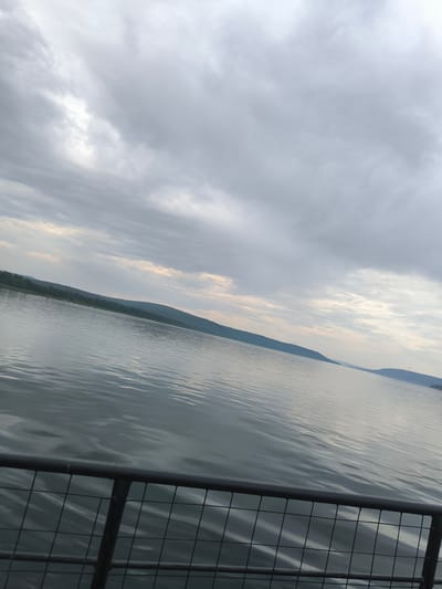 The image is an exterior, eye-level shot, taken from a slightly tilted perspective, of a large body of water, likely a river, bordered by forested hills under a cloudy sky. In the foreground, a dark metal railing with a wire mesh fence is visible, suggesting the viewpoint is from a dock, pier, or boat. The water is calm with gentle ripples reflecting the muted light of the overcast sky. The distant shoreline features rolling hills covered in dense green trees, with some subtle variations in elevation. The sky is filled with a thick layer of grey clouds, with some lighter patches indicating diffused sunlight breaking through. The overall impression is one of a natural landscape on a somewhat gloomy day. The location is identified as Boguchany, Russia, a settlement situated on the Angara River, which fits the visual evidence of a wide river and surrounding hills.