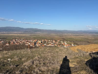 The image presents a panoramic, high-angle view overlooking the village of Novo Leski, Bulgaria, and its surrounding environment under clear daylight. The foreground consists of dry, unkempt grass and sparse, brown vegetation covering an uneven hillside. A distinct, elongated human shadow, cast by the photographer, extends across the lower-right quadrant of this foreground.
The midground is dominated by the village itself, which comprises numerous residential buildings, largely characterized by orange-red tiled roofs. These structures are densely clustered, interspersed with dormant trees and shrubs. Beyond the village, a wide, flat valley stretches into the distance, appearing as a patchwork of brown and green agricultural fields and undeveloped land.
The background is defined by a range of moderate-sized mountains with visible tree cover, forming the distant horizon. The sky above is bright, clear blue with a few wispy white clouds scattered across the upper portions. The scene is illuminated by direct sunlight, indicated by the prominent shadow in the foreground. No other individuals or specific actions are discernible within the visible landscape.
