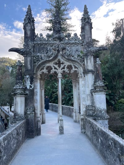 An intricately carved stone structure dominates the frame. Its Gothic architecture is emphasized by pointed arches, ornate detailing, and lichen-covered surfaces, suggesting age and weathering. A figure in dark clothing stands within the structure's open space. Lush greenery peeks through the openings and surrounds the site. The sky, with scattered clouds, forms a backdrop. The scene conveys a sense of history, architecture, and peaceful solitude.