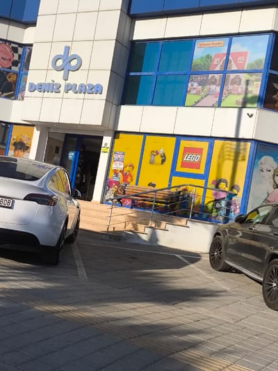 The image displays the exterior of Deniz Plaza, a commercial building located in Gonyeli, Northern Cyprus. The building features a multi-story, white-tiled facade with blue-tinted windows. A prominent "dp" logo and "DENIZ PLAZA" signage are affixed to the upper section of the building.

The ground floor of the building consists of retail window displays. To the left of the main entrance, a window shows a child's face and various backpacks. The central and right sections of the ground floor feature multiple windows with bright yellow backgrounds showcasing "LEGO" branding and Lego minifigure displays. An adjacent window on the far right displays characters from Disney's Frozen. On the first floor, some windows are blue-tinted, while others display "Sylvanian Families" merchandise, including miniature houses and animal figures.

The main entrance to Deniz Plaza is recessed and accessed via a short flight of concrete steps, accompanied by a silver-colored metal handrail. A small white sign bearing the Turkish word "ÇIK" (Exit) is visible near the entrance.

Two vehicles are parked on the grey paved forecourt in front of the building. On the left, a white Tesla Model Y is partially visible from the rear, with its license plate partially obscured, showing "BDB 808". On the right, a dark-colored Mercedes-Benz SUV is partially visible from its front-side aspect. The forecourt features white parking space markings and tactile paving strips. Direct sunlight illuminates the scene, casting distinct shadows across the paved area. No individuals are present in the image.