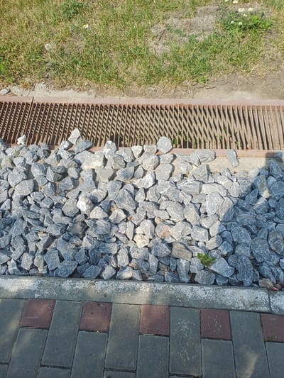 This is a top-down view of an outdoor scene. The image is divided into sections showcasing different ground textures. On the left is a patch of green grass with a few small white flowers. Adjacent to the grass is a metal grate, likely a drain cover, with parallel slats. To the right of the grate is an area covered with numerous gray, angular rocks. Further to the right, a border of concrete separates the rocks from a paved walkway made of rectangular bricks, alternating in color between gray and reddish-brown.
There are no people or animals visible in the image. The setting appears to be a park, garden, or public pathway. The lighting suggests it is daytime, with shadows cast by the grate and some of the rocks. The weather appears to be clear and sunny. The rocks and paving stones suggest an urban or suburban outdoor environment. The presence of grass and flowers indicates a natural element within this setting. There is no visible text in the image.